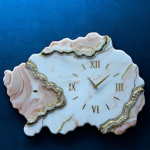 May include: White and pink resin clock with gold Roman numeral markings and a gold-toned hour and minute hand. The clock is shaped like an irregular, organic shape with a white background and pink and gold accents.