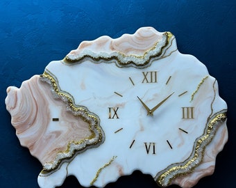 May include: White and pink resin clock with gold Roman numeral markings and a gold-toned hour and minute hand. The clock is shaped like an irregular, organic shape with a white background and pink and gold accents.