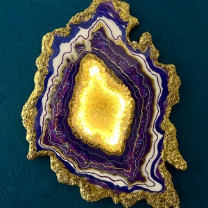 May include: A decorative, abstract art piece resembling a geode. The artwork features concentric layers of white, purple, and gold, with a textured gold border. The center has a bright, golden glow, set against a teal background.