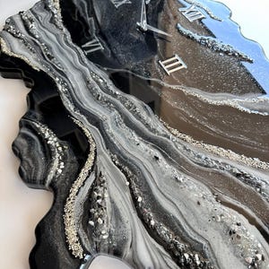 May include: A wall clock with an irregular, organic shape featuring a glossy resin finish in black, white, and gray swirls. The surface is accented with silver glitter and crushed glass textures. It includes silver Roman numeral markers and hands.