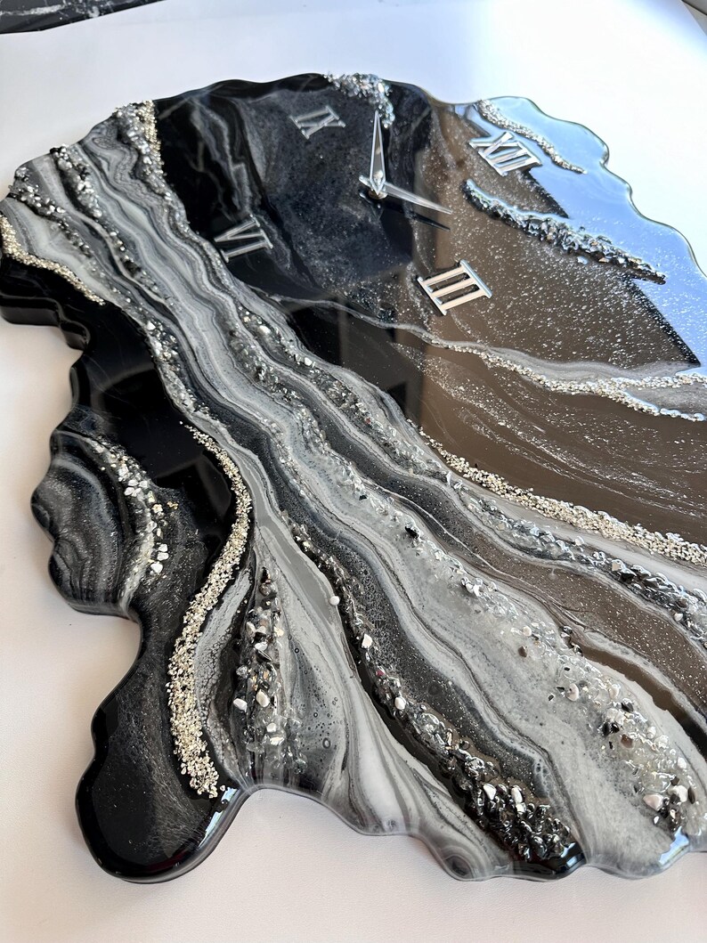 May include: A wall clock with an irregular, organic shape featuring a glossy resin finish in black, white, and gray swirls. The surface is accented with silver glitter and crushed glass textures. It includes silver Roman numeral markers and hands.