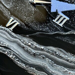 May include: A wall clock with a glossy black resin surface featuring swirling white and gray marbled patterns. The clock face includes silver Roman numerals for XII, III, VI, and IX, along with silver clock hands set against a glittery finish.