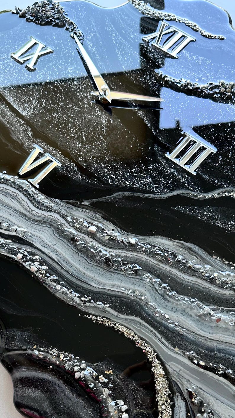 May include: A wall clock with a glossy black resin surface featuring swirling white and gray marbled patterns. The clock face includes silver Roman numerals for XII, III, VI, and IX, along with silver clock hands set against a glittery finish.