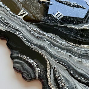 May include: A wall clock with an irregular, organic shape featuring a glossy black, white, and silver marbled resin finish. The clock face includes silver Roman numerals at the three, six, nine, and twelve o'clock positions and silver hands.