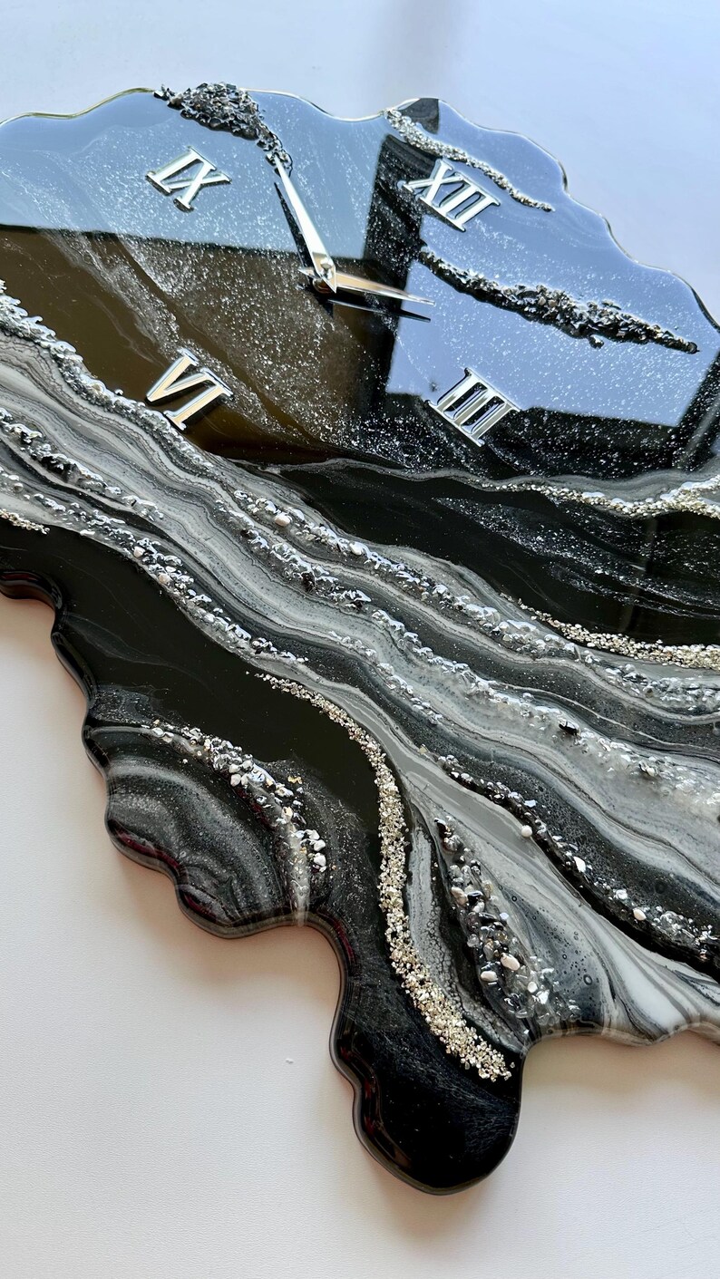 May include: A wall clock with an irregular, organic shape featuring a glossy black, white, and silver marbled resin finish. The clock face includes silver Roman numerals at the three, six, nine, and twelve o'clock positions and silver hands.