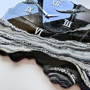 May include: A wall clock with an irregular, organic shape featuring a glossy black, white, and silver resin finish. The surface displays swirling marbled patterns and textured metallic accents. It includes silver clock hands and Roman numerals XII, III, VI, and IX.