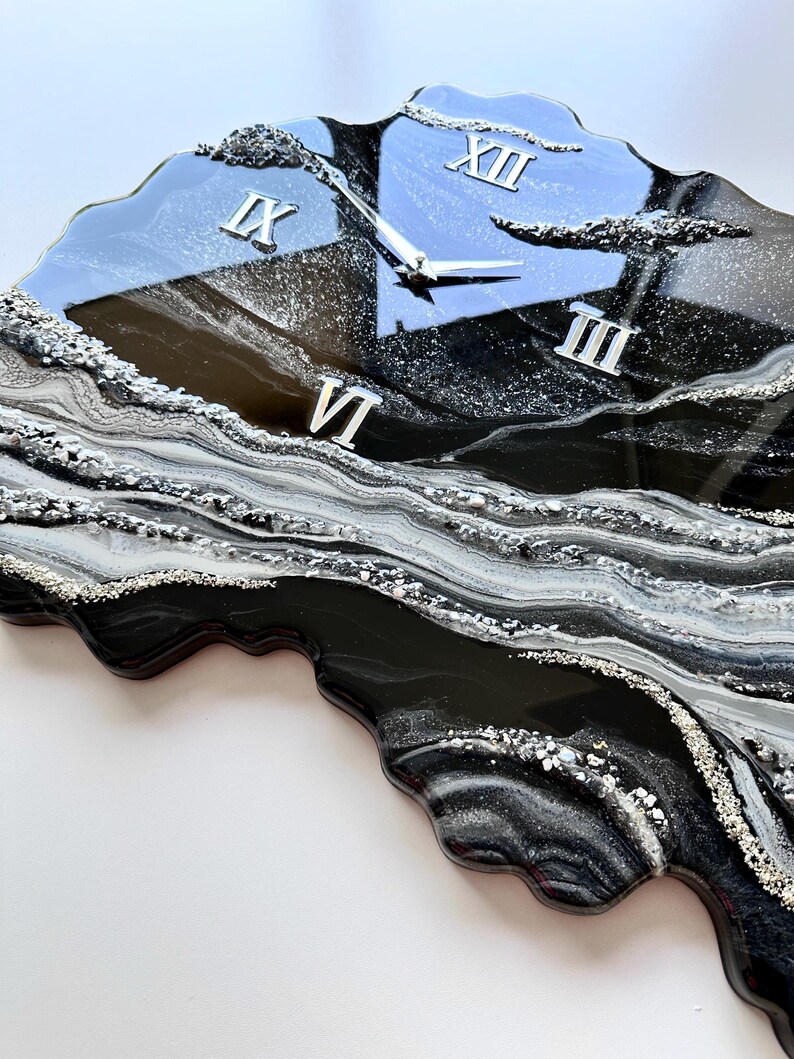May include: A wall clock with an irregular, organic shape featuring a glossy black, white, and silver resin finish. The surface displays swirling marbled patterns and textured metallic accents. It includes silver clock hands and Roman numerals XII, III, VI, and IX.