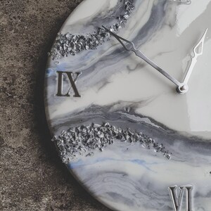 May include: A white and gray resin clock face with silver glitter embedded in the resin. The clock face has Roman numerals for the hours, and the hands are silver. The clock face is round and has a white background with gray and blue swirls.