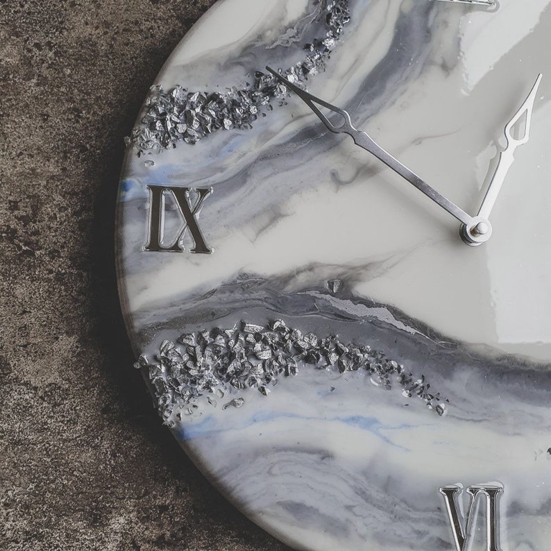 May include: A white and gray resin clock face with silver glitter embedded in the resin. The clock face has Roman numerals for the hours, and the hands are silver. The clock face is round and has a white background with gray and blue swirls.