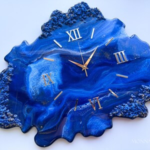 May include: Blue and gold resin wall clock with Roman numeral markings. The clock face is an abstract design with a blue and white marbled effect.