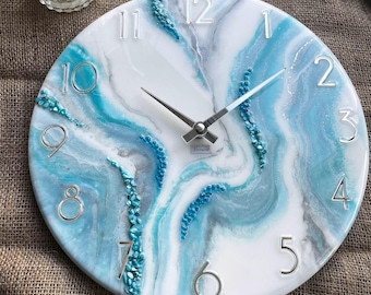 May include: Round wall clock with a white face, featuring a blue and gray marbled design. Silver numerals mark the hours, and the clock hands are silver. Decorative blue stones accent the design. The clock is a decorative home decor item.