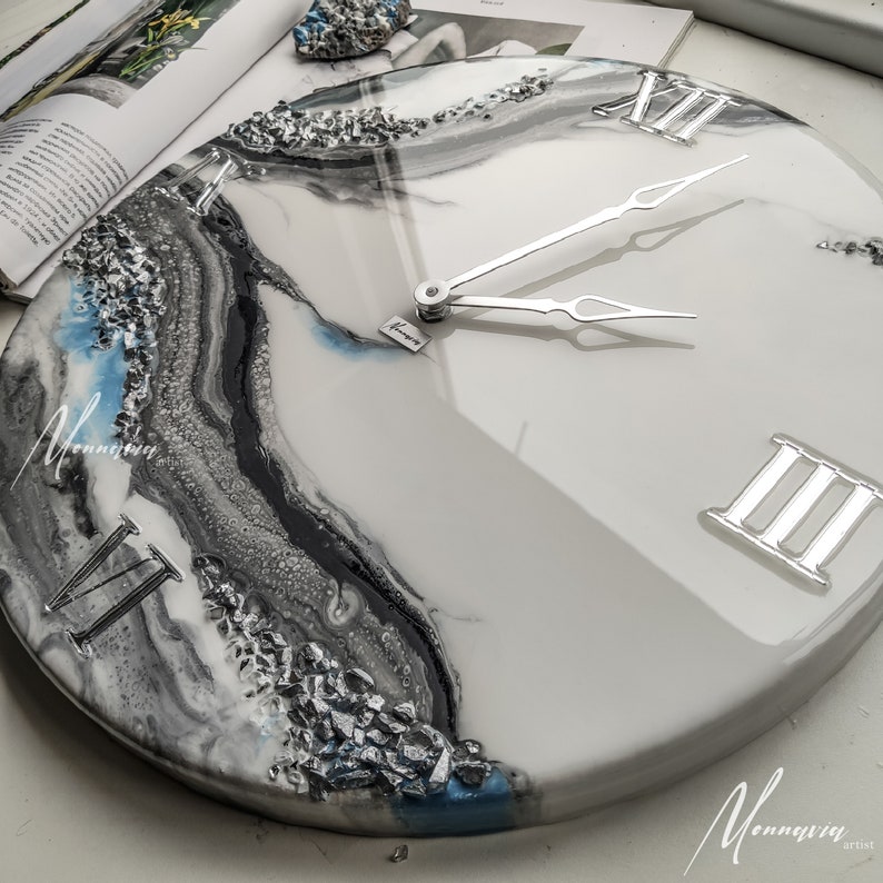 May include: A white resin clock with silver Roman numeral hour markers and silver hands. The clock face has a blue, black, and silver abstract design.