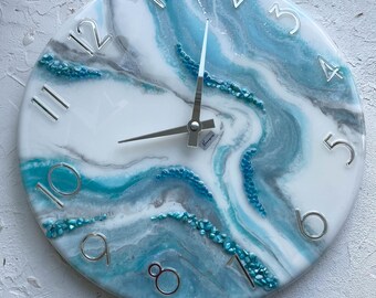 May include: A round, white clock face with blue and gray abstract swirls. The clock face has silver Roman numeral hour markers and silver hands. The clock is made of resin and has a unique, modern design.