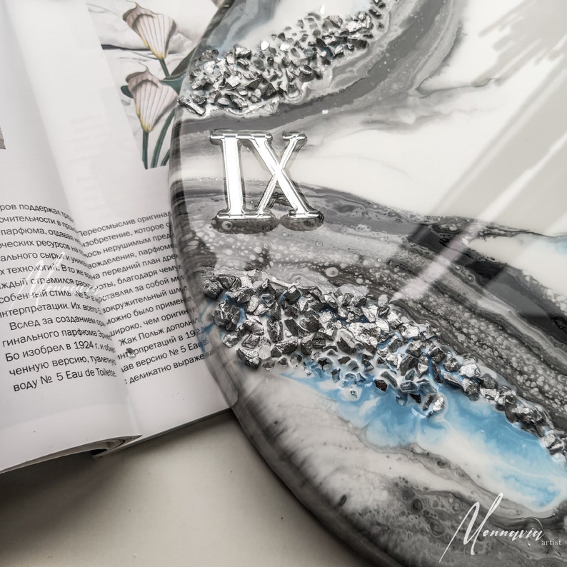 May include: A close-up of a round, white, gray, and blue resin clock face with silver glitter embedded in the resin. The clock face has Roman numerals, with the number IX visible in the image. The clock face is on a white surface.
