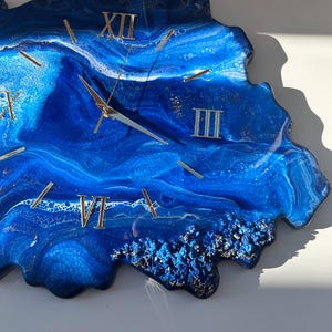 May include: Blue resin clock with gold Roman numeral markings and a white background. The clock face is shaped like a wave.