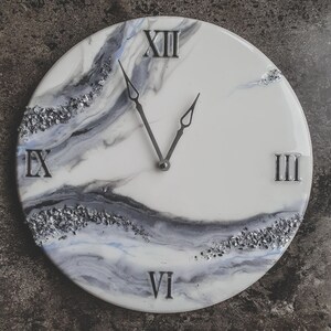 May include: A white and gray resin clock with Roman numeral markings. The clock face has a marbled design with silver glitter accents.