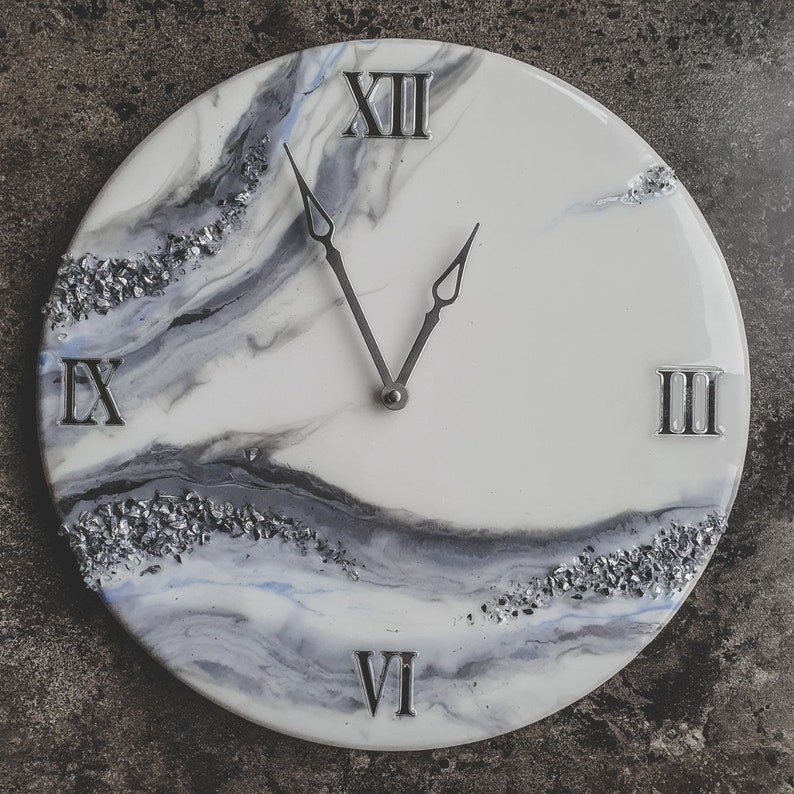May include: A white and gray resin clock with Roman numeral markings. The clock face has a marbled design with silver glitter accents.