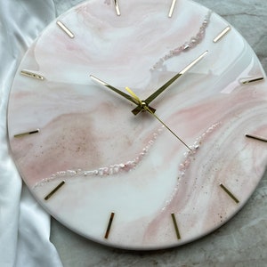 May include: A white and pink resin clock with gold hands and gold hour markers. The clock face has a marbled design with embedded glitter and small white stones.