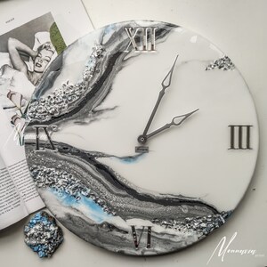 May include: A white, gray, and blue resin clock with Roman numeral markings and silver hands. The clock face has a marbled design with silver glitter accents.