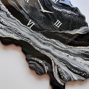 May include: An irregular, geode-inspired wall clock featuring a glossy black, white, and silver marbled resin finish. The clock face displays silver Roman numerals at the three, six, nine, and twelve o'clock positions with matching silver hands.