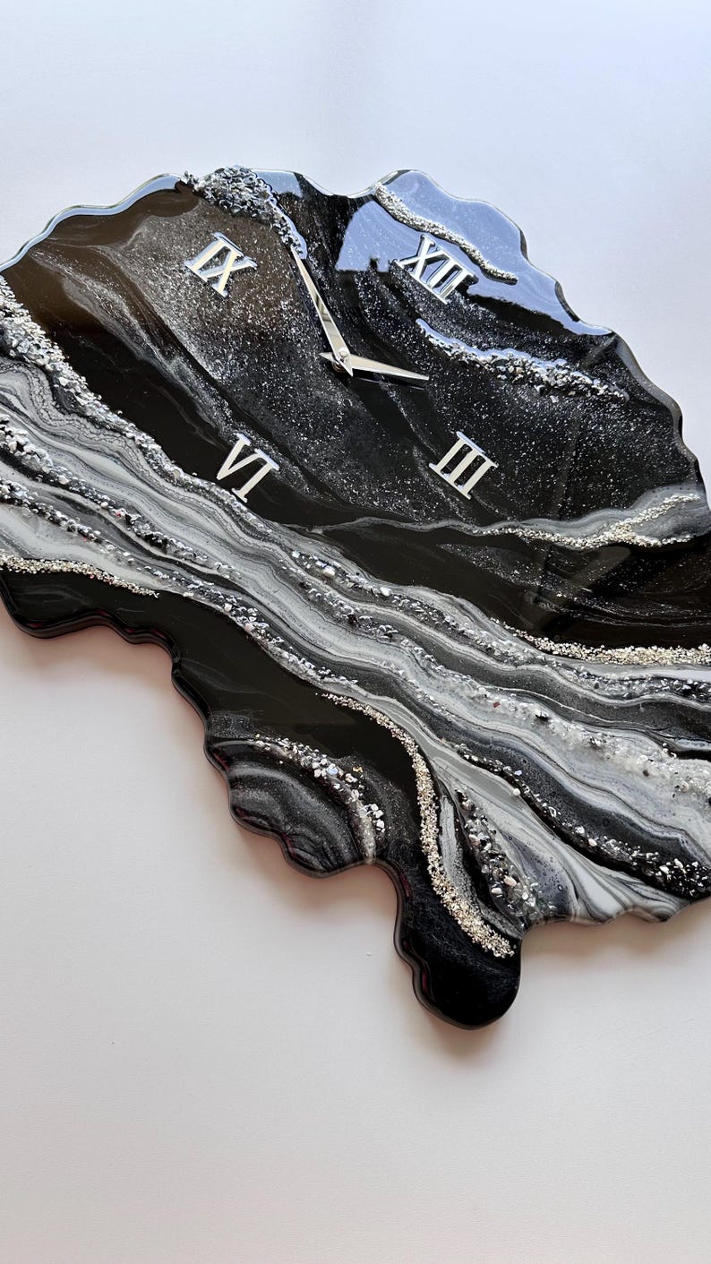 May include: An irregular, geode-inspired wall clock featuring a glossy black, white, and silver marbled resin finish. The clock face displays silver Roman numerals at the three, six, nine, and twelve o'clock positions with matching silver hands.