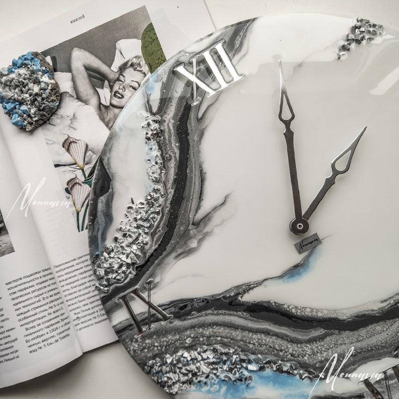 May include: A white resin clock with black and silver abstract swirls. The clock face has Roman numerals and silver hands. The clock is 12 inches in diameter.