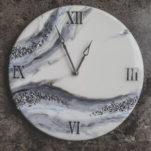 May include: A white clock face with black and gray marbling and silver glitter accents. The clock face has Roman numerals and silver hands. The clock is round and has a white background.