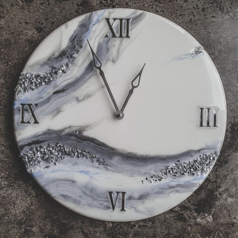 May include: A white clock face with black and gray marbling and silver glitter accents. The clock face has Roman numerals and silver hands. The clock is round and has a white background.