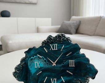 May include: A teal and silver resin clock with Roman numerals. The clock has a unique, organic shape and sits on a white table. The background includes a white sofa and a painting.