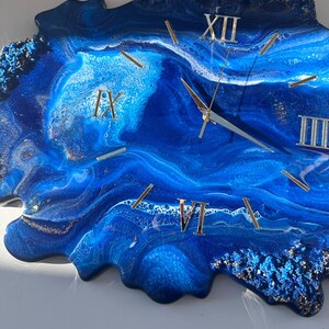 May include: Blue and white resin clock with gold Roman numeral markings. The clock face is a unique abstract design with a blue and white swirl pattern.