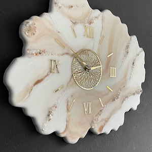 May include: A white and beige resin clock with gold Roman numeral hour markers and a gold metal clock face. The clock is shaped like a flower and has small clear crystals embedded in the resin.