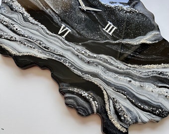 May include: A wall clock with an irregular, organic shape featuring a glossy black, white, and silver marbled finish. The clock face includes silver Roman numerals for three, six, nine, and twelve, along with silver clock hands and textured accents.