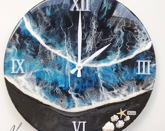 Puede incluir: Un reloj redondo de resina con un diseño de ola de océano azul y blanco. La esfera del reloj tiene números romanos y manecillas blancas. El reloj está decorado con conchas marinas y una estrella de mar.