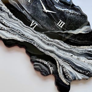 May include: An irregular, geode-inspired wall clock featuring a glossy black, white, and silver resin finish. The surface displays swirling marbled patterns with embedded silver glitter accents. It includes silver clock hands and Roman numerals.