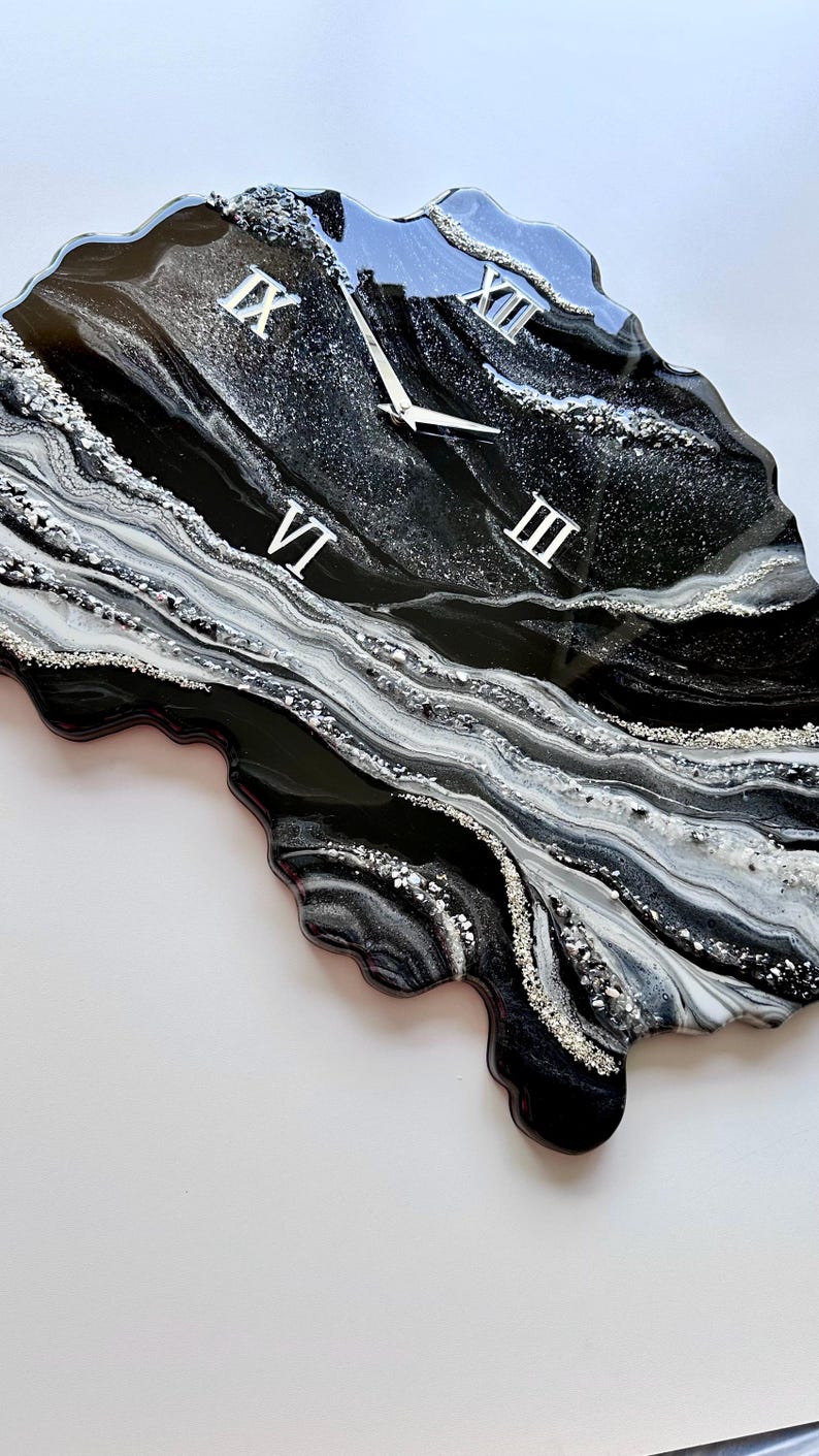 May include: An irregular, geode-inspired wall clock featuring a glossy black, white, and silver resin finish. The surface displays swirling marbled patterns with embedded silver glitter accents. It includes silver clock hands and Roman numerals.