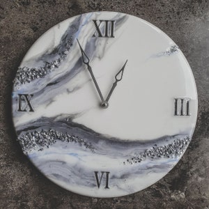 May include: A white and gray resin clock with Roman numeral markings. The clock face has a marbled design with silver glitter accents.