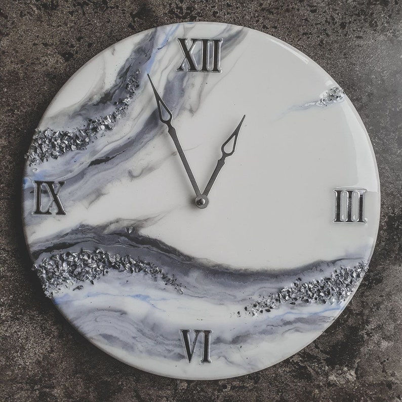 May include: A white and gray resin clock with Roman numeral markings. The clock face has a marbled design with silver glitter accents.
