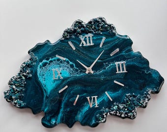 May include: A teal and dark blue resin wall clock with silver Roman numerals. The clock has a unique, organic shape with a textured, glittery border. The clock hands are silver. The clock face has a swirling, marbled effect.