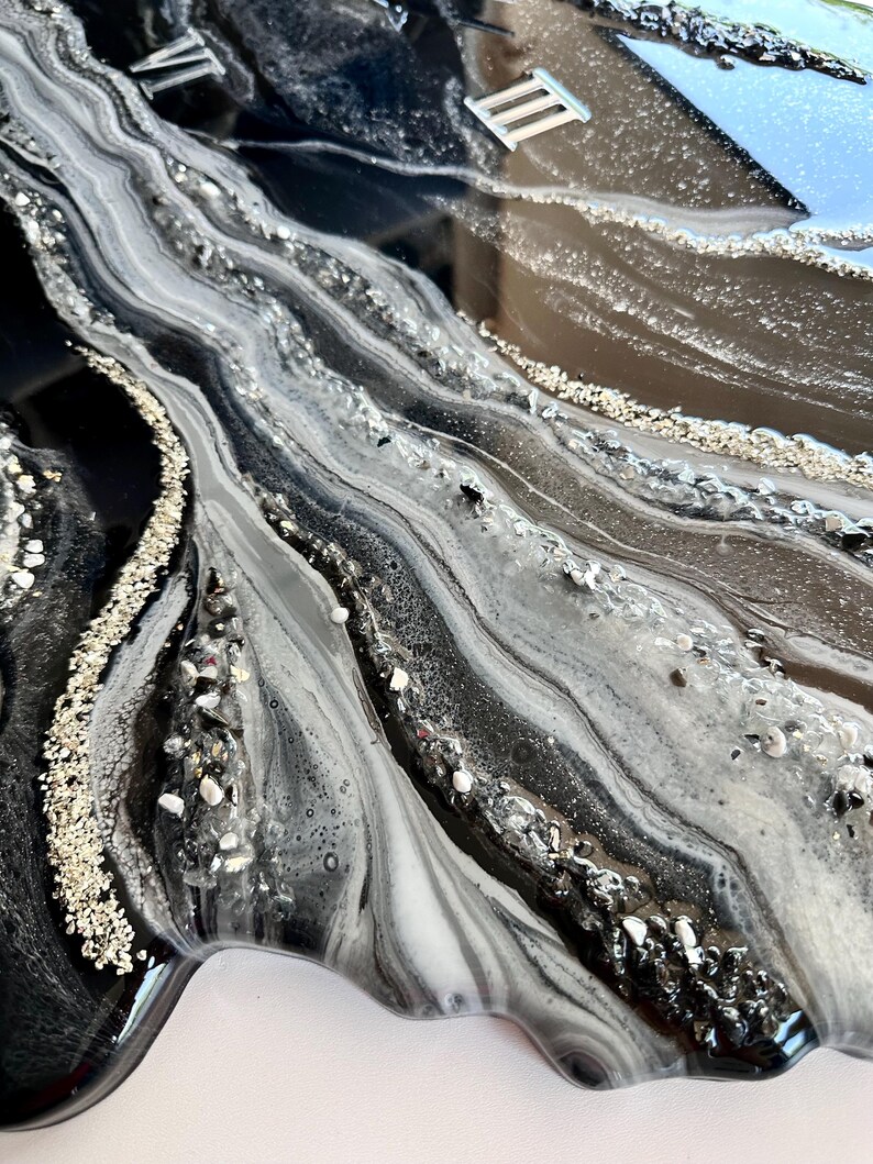 May include: A wall clock featuring a glossy black and white marbled resin surface with flowing, wave-like patterns. The design incorporates embedded silver-toned crushed glass and metallic accents, with silver Roman numerals III and VI visible.