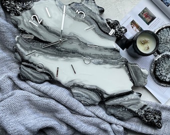 May include: A decorative wall clock with a unique, organic shape. The clock face features a marbled design in shades of gray and white, with silver-colored hands and hour markers. The clock is resting on a gray knitted blanket.