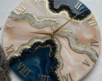 May include: A round clock with Roman numeral markings on a white and blue resin background. The clock face features a swirling, abstract design with gold accents and black and white glitter.