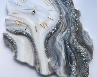 May include: A decorative wall clock with a unique, organic shape. The clock face features a marbled design in shades of white, gray, and silver, with gold Roman numerals and hands. The edges are embellished with sparkling crystals.