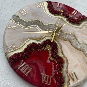 Könnte beinhalten: Eine runde Uhr aus weißem und rotem Harz mit goldenen römischen Ziffern und goldenen Zeigern. Das Zifferblatt hat ein wirbelndes, achatähnliches Design mit goldenen und weißen Glitzerakzenten.