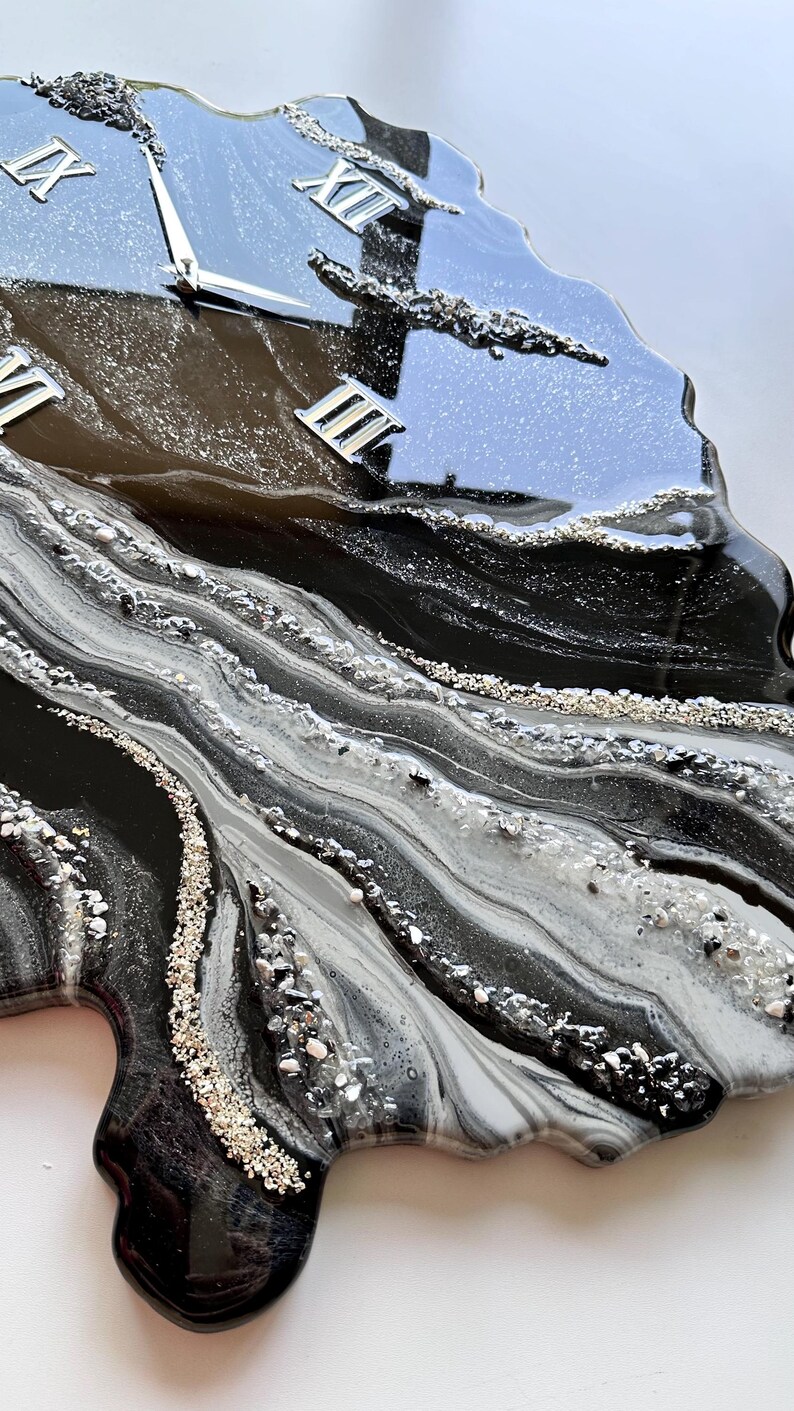 May include: A wall clock with an irregular, organic shape featuring a glossy black, white, and gray marbled resin finish. The surface is accented with silver glitter and crushed glass textures. It includes silver Roman numerals and clock hands.