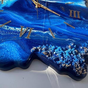 May include: A blue and gold resin clock with Roman numeral markings. The clock face is a swirling blue and white abstract design with gold glitter accents.