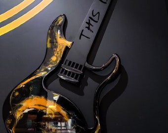 May include: Black electric guitar with gold and black marbling on the body. The guitar has a black neck and a black pickguard. The text "This is" is written on the neck of the guitar.