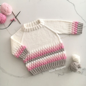 May include: A white knitted baby sweater with pink and gray stripes. The sweater has a round neckline and long sleeves. The sleeves have a textured pattern.