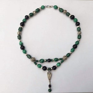 Op de afbeelding: Een ketting met twee strengen met groene en zwarte kralen, afgewisseld met zilverkleurige decoratieve kralen. De ketting heeft een zilverkleurige hanger met een bungelende kraal. De ketting wordt op een wit oppervlak getoond.