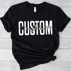 Op de afbeelding: Zwart T-shirt met witte tekst die "CUSTOM custom" in een gestileerd lettertype luidt.