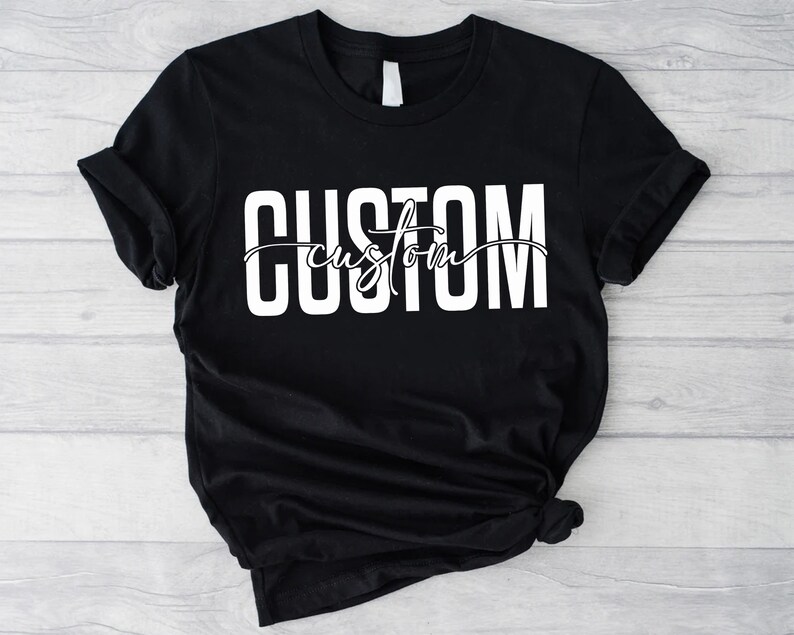 Op de afbeelding: Zwart T-shirt met witte tekst die "CUSTOM custom" in een gestileerd lettertype luidt.